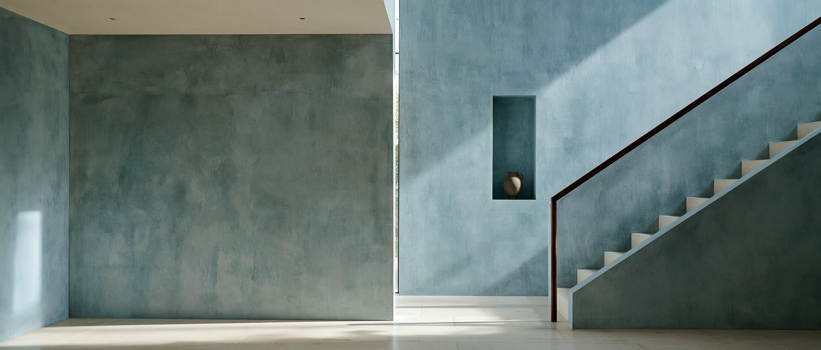 A blue-gray limewashed hallway with a wall niche and a plaster staircase with a dark wooden handrail